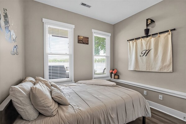A bedroom with a bed, two windows with blinds, a wall-mounted towel rack with a towel, and a small shelf with flowers.