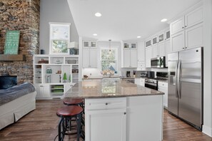 A modern kitchen with white cabinetry, a large island with stools, a stone accent wall, and a view of the outdoors.