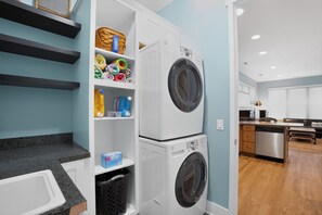 A laundry room with stacked washers and dryers, shelves with laundry supplies, and a kitchen area in the background.
