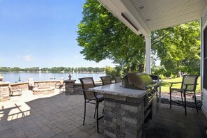 An outdoor patio with a stone fire pit, seating, and a grill, overlooking a lake.