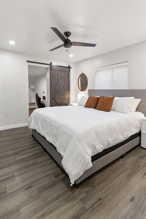 A bedroom with a large bed, a ceiling fan, a wooden door, a window with blinds, and a small desk with a chair.