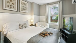 Un dormitorio ordenado con cama, mesita de noche con lámpara, una silla, ventana con cortinas y televisor.