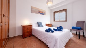 Un dormitorio con cama, mesita de noche, silla, ventana y un cuadro en la pared.