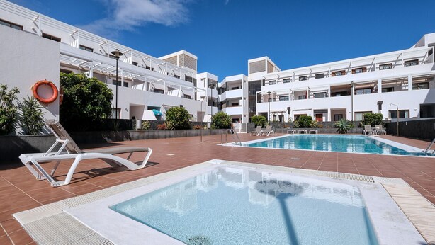 Zona de piscina con dos piscinas, sillas de descanso y un edificio blanco con balcones.