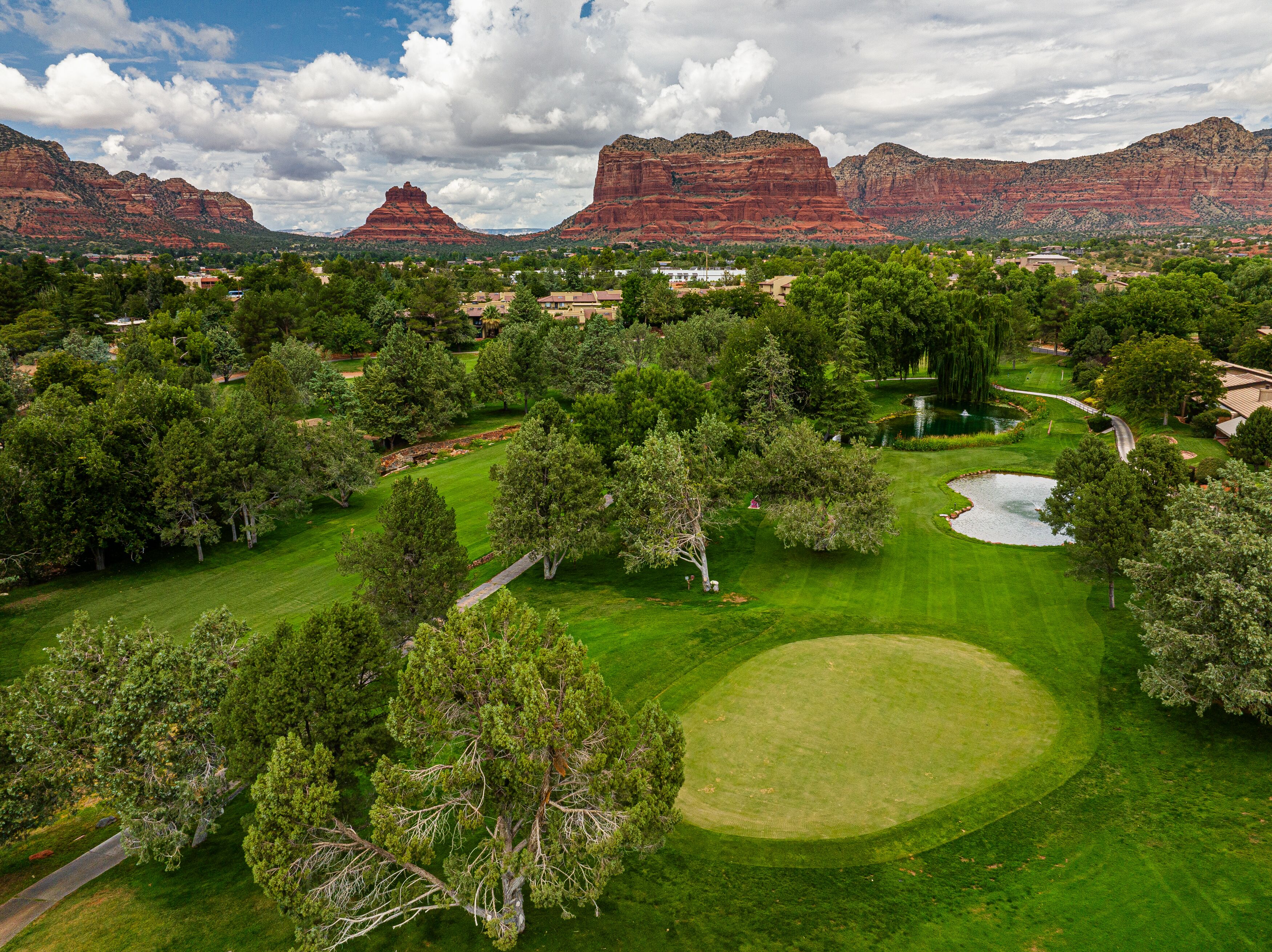 Un campo de golf con un estanque, rodeado de árboles y una cadena montañosa al fondo.