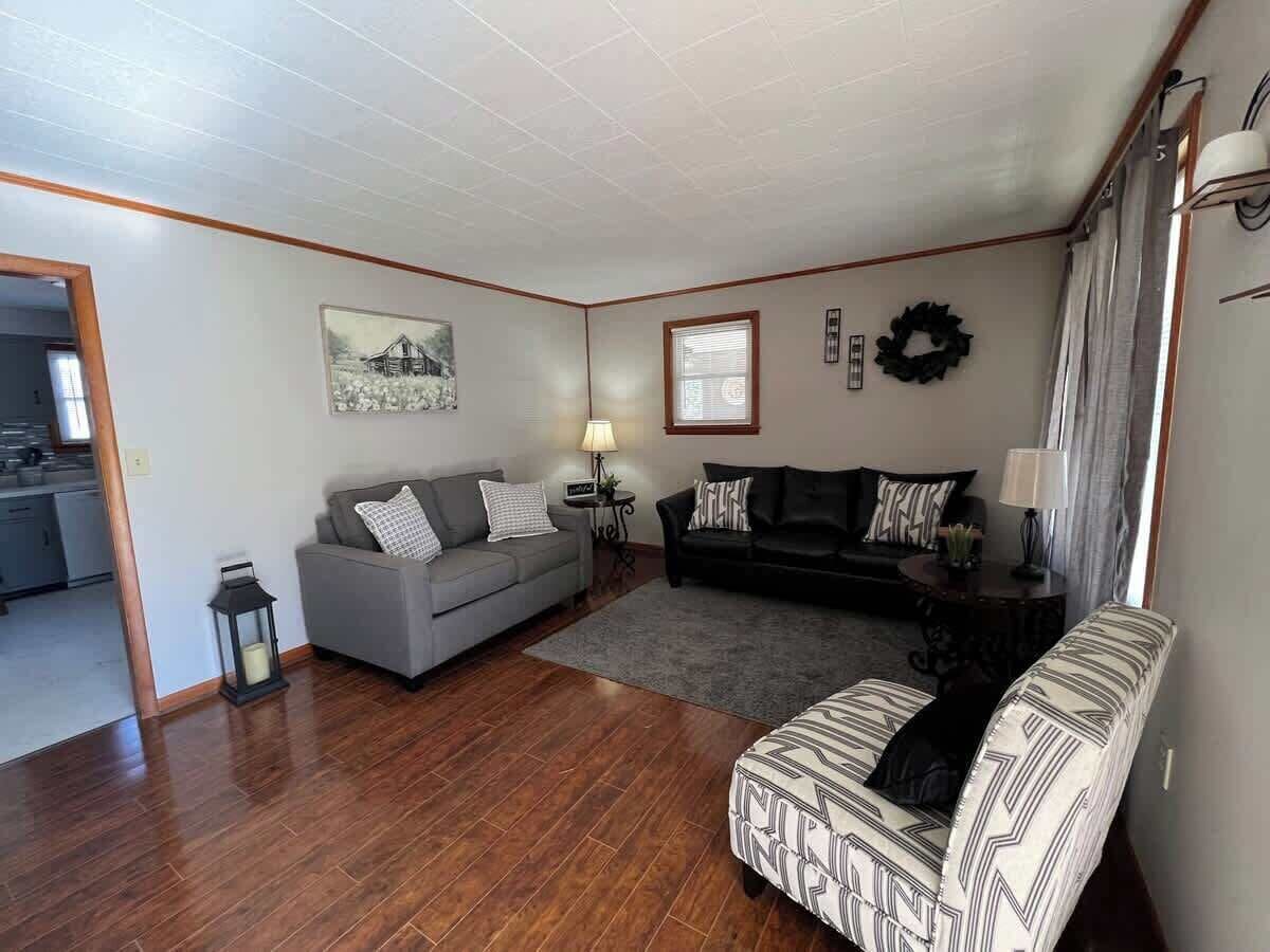 Una sala de estar con un sofá gris, un sofá negro, un sillón estampado, piso de madera e imagen colgada en la pared.
