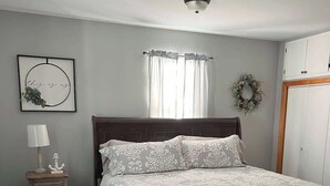 A bedroom with a bed, a nightstand, a wall-mounted lamp, a window with curtains, a wall decoration, and a wardrobe.