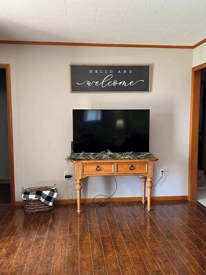 Quarto com piso de madeira, uma TV de tela plana sobre um console de madeira, e uma placa de boas-vindas acima da TV.