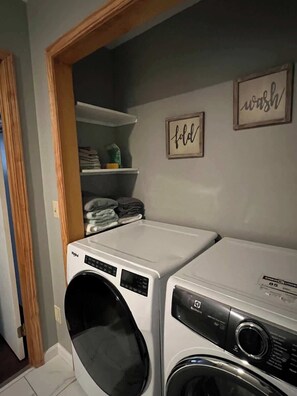 A laundry room with two washing machines, a shelf with folded clothes, and two signs on the wall.