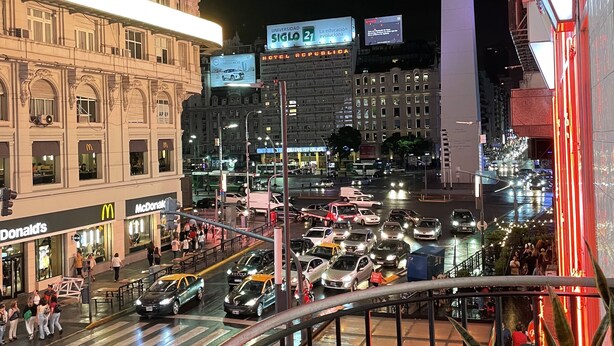 Affollata via cittadina di notte, con edifici illuminati, un McDonald's e l'Hotel Republica.