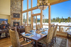 A dining area with a stone fireplace, wooden furniture, and large windows offering a view of a snowy landscape. - Moose Run Spacious Winter Park Area Retreat (Fraser)