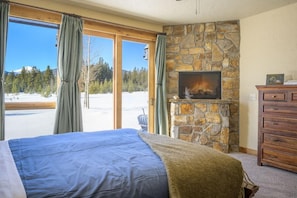 Une chambre dotée d’un grand lit, d’une cheminée en pierre, d’une commode et offrant une vue sur un paysage enneigé grâce à de grandes fenêtres.