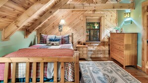 A cozy attic bedroom with a wooden ceiling, a bed with plaid bedding, a nightstand, a dresser, and a window with a view of greenery.