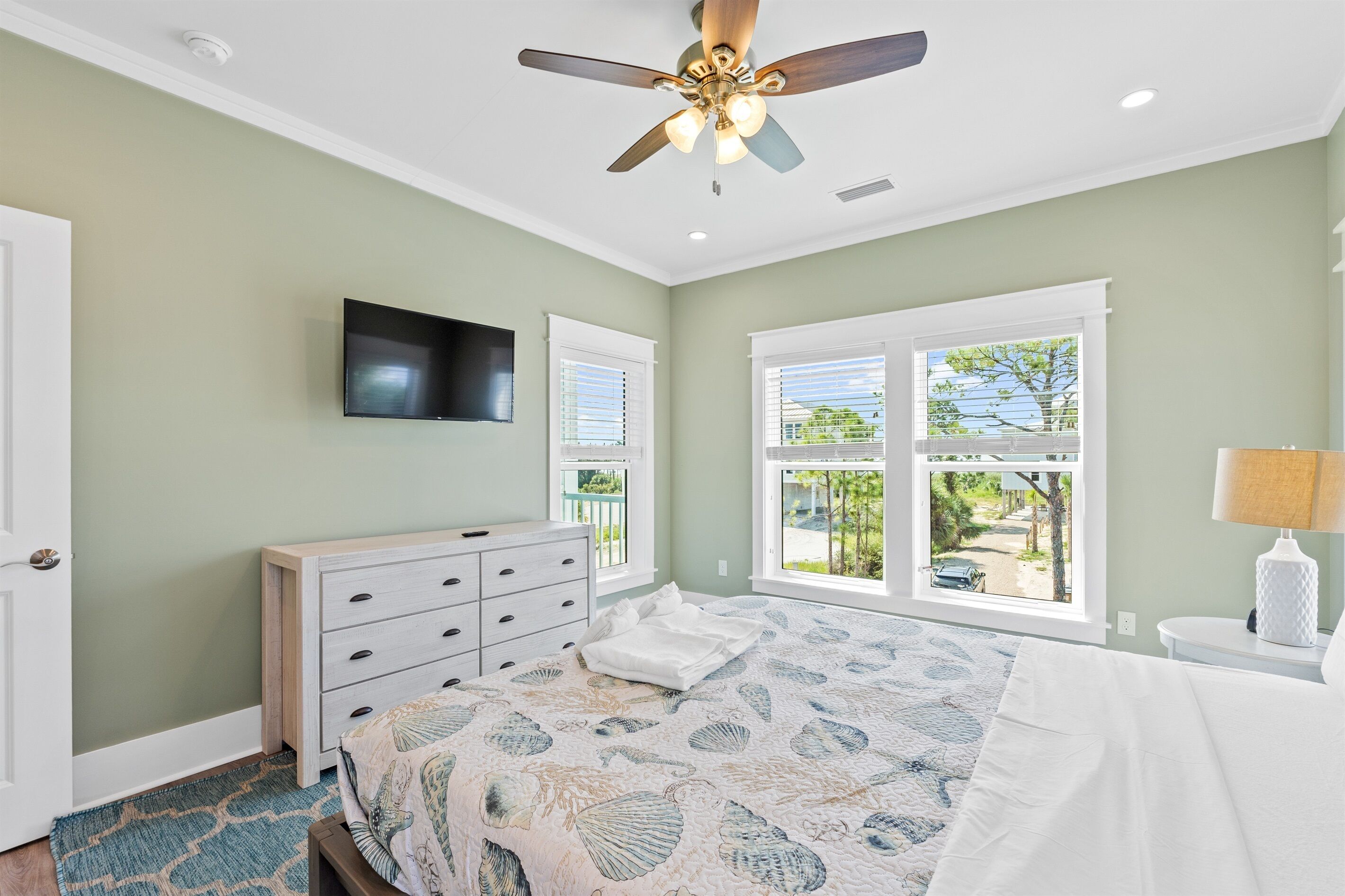 A bedroom with a bed, a dresser, a ceiling fan, a TV, and a window with a view.