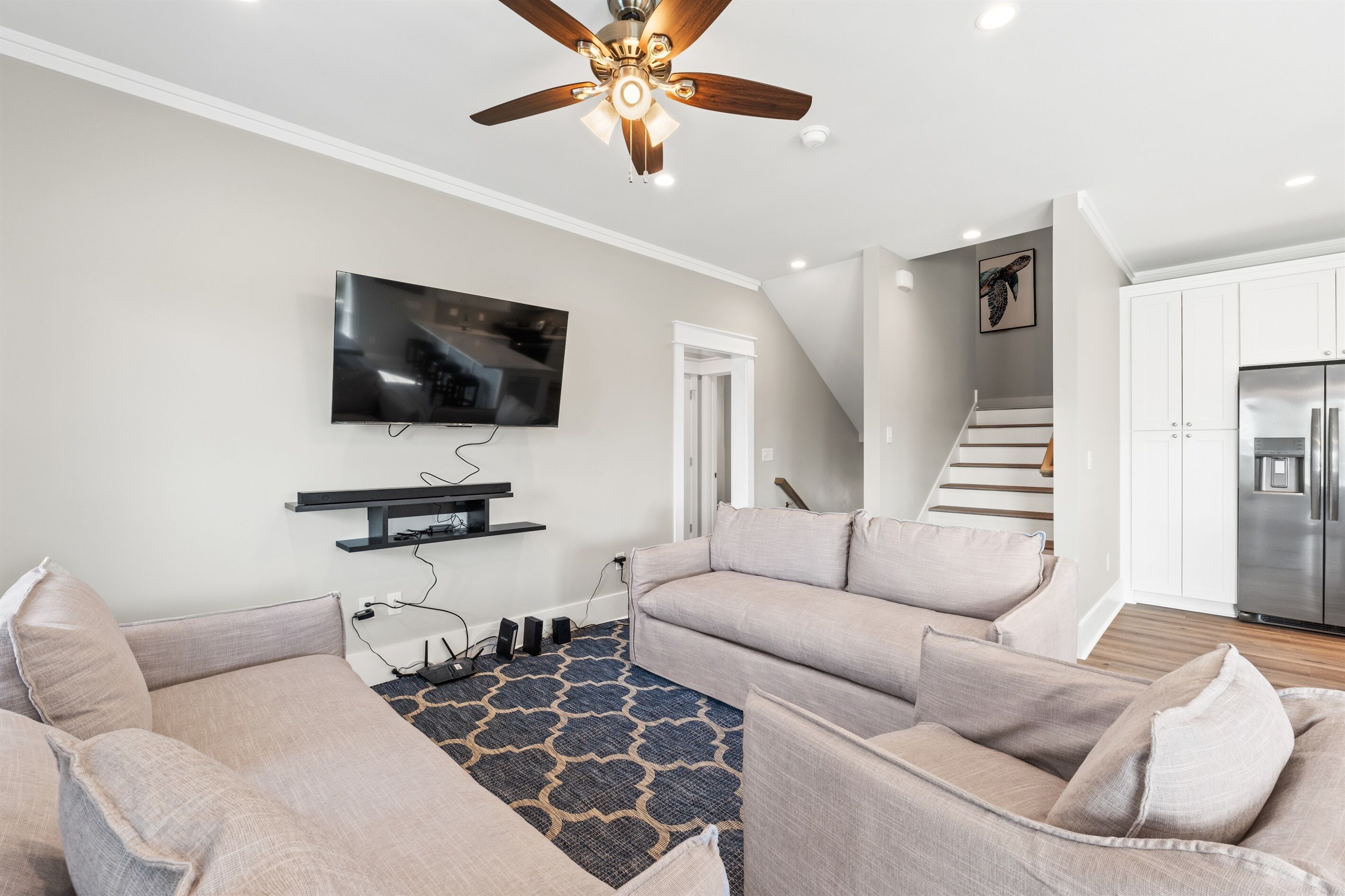 A modern living room with a flat-screen TV, ceiling fan, and a staircase leading to a kitchen.