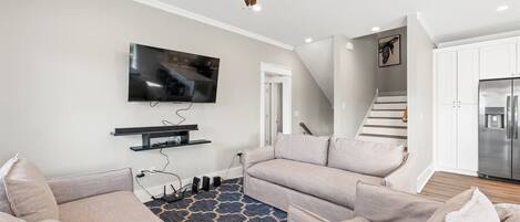 A modern living room with a flat-screen TV, ceiling fan, and a staircase leading to a kitchen.