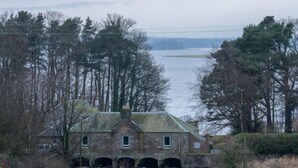 Un bâtiment en pierre avec un pont enjambant une rivière, entouré d’arbres et avec une étendue d’eau en arrière-plan.