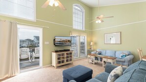 Un salon comprenant un canapé, un pouf et un téléviseur posé sur un meuble en bois. On y trouve une fenêtre avec des stores, un ventilateur de plafond et une porte donnant sur un balcon.