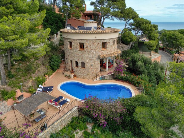 A stone villa with a swimming pool, surrounded by lush greenery and overlooking the sea.