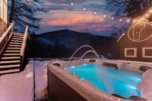 Un bain à remous avec un éclairage bleu, entouré de neige, avec un escalier et un bâtiment dont le toit est surmonté de la silhouette d’une personne.
