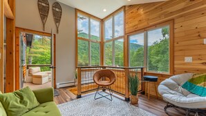 A cozy room with a green sofa, a white chair, and a view of lush greenery through large windows. - Secluded/Hot Tub/views! Minutes to Everything! (Bridgewater Corners)