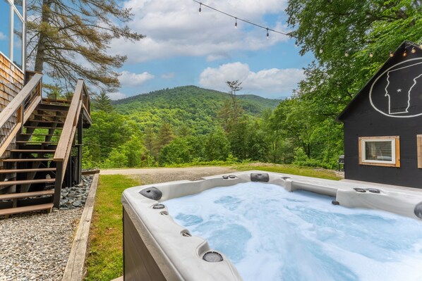 Un bain à remous doté d’une terrasse en bois et d’un escalier, entouré d’un paysage verdoyant luxuriant et avec une montagne en arrière-plan.