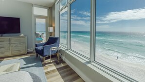 Een kamer met een groot raam met uitzicht op een strand, een televisie, een stoel en een klein tafeltje.