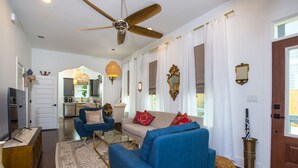 A spacious living room with a blue sofa, a beige sofa, and a glass coffee table. There is a ceiling fan, a wooden cabinet, and a decorative mirror on the wall.