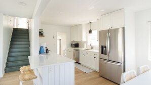 Une cuisine moderne avec des meubles blancs, des appareils en acier inoxydable, un îlot central doté de tabourets et un escalier menant à un autre niveau.