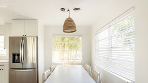 Une cuisine moderne avec des meubles blancs, un réfrigérateur en acier inoxydable, une table à manger avec six chaises et une grande fenêtre équipée de stores.