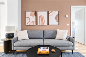 A modern living room with a grey sofa, two white cushions, a black coffee table with a magazine and a small stack of books, a lamp, and abstract wall art.
