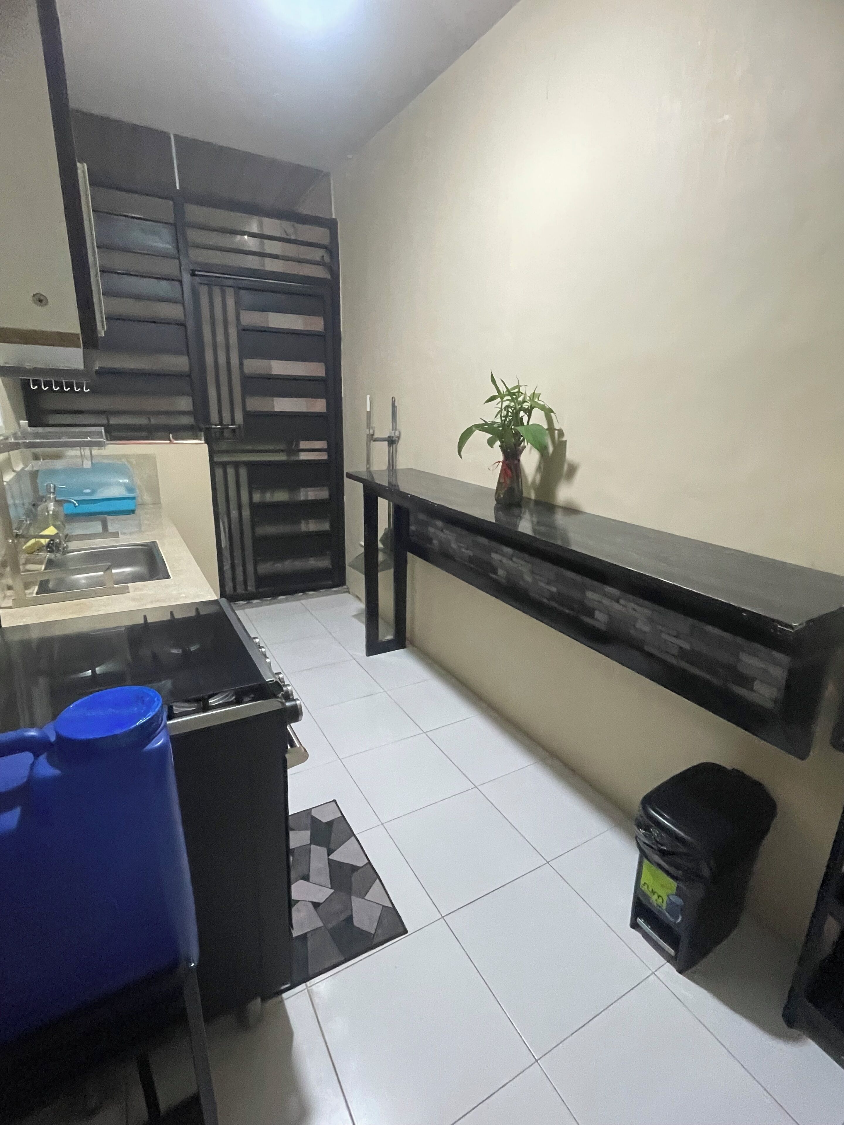 Una cocina con estufa a gas, lavamanos, sillas azules, una planta en maceta y un cubo de basura negro.