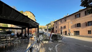An outdoor café with tables and chairs, a covered seating area, and a street view with buildings and a clear sky.