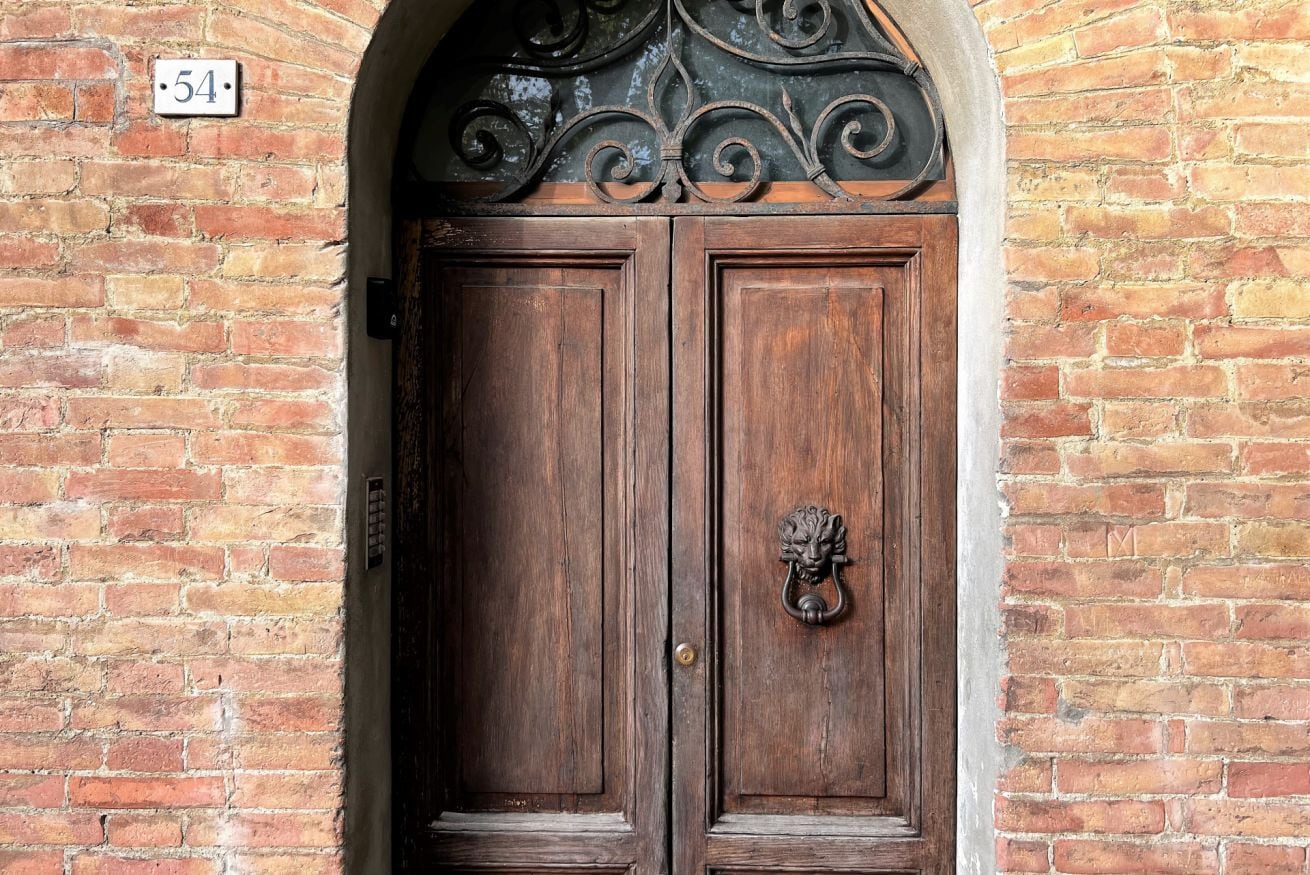 Una puerta doble de madera con un llamador con forma de cabeza de león, ubicada en un muro de ladrillo con un cartel con el número 54.