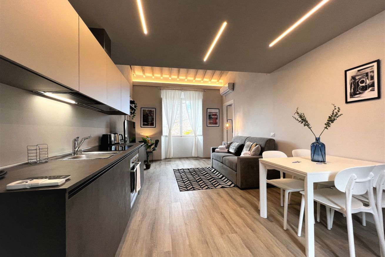 Una cucina moderna con isola centrale, una zona pranzo con tavolo e sedie bianche, e un angolo living con divano e tavolino.