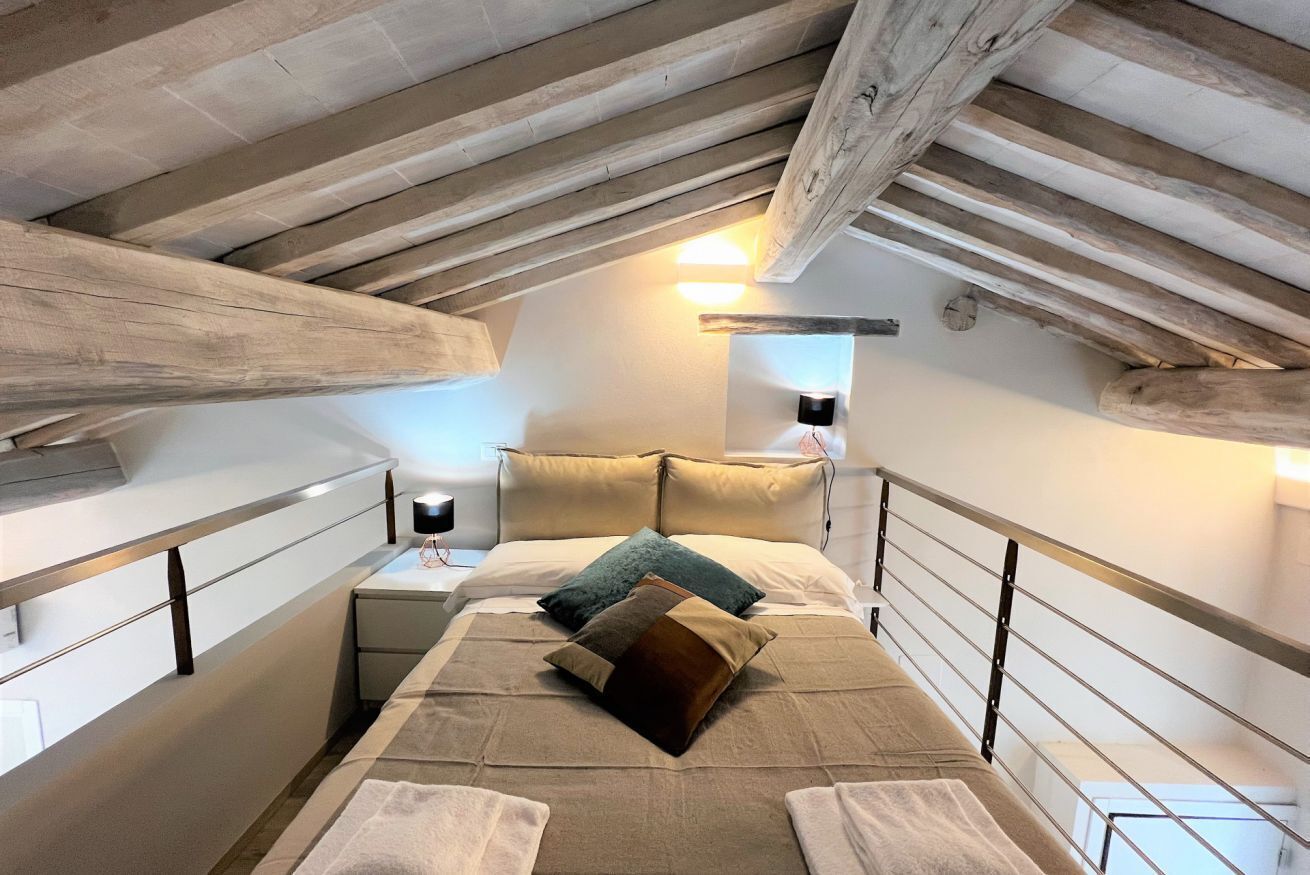 Ein Schlafzimmer im Loft-Stil mit schräg verlaufender Decke, freiliegenden Holzbalken, einem Bett mit vielen Kissen, Nachttischen mit Lampen und einem weißen Kleiderschrank.