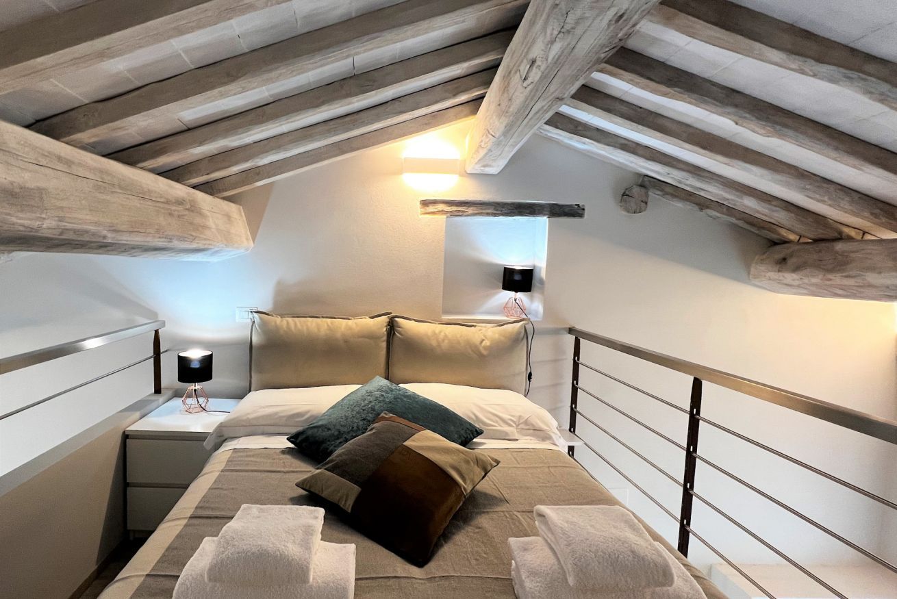 Un dormitorio con techo inclinado, una cama con varias almohadas, una mesita de noche con lámpara y una lámpara de pared.