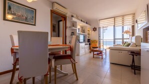 A modern living room with a dining area, a sofa, and a TV. There is a large window with a view of the sea.