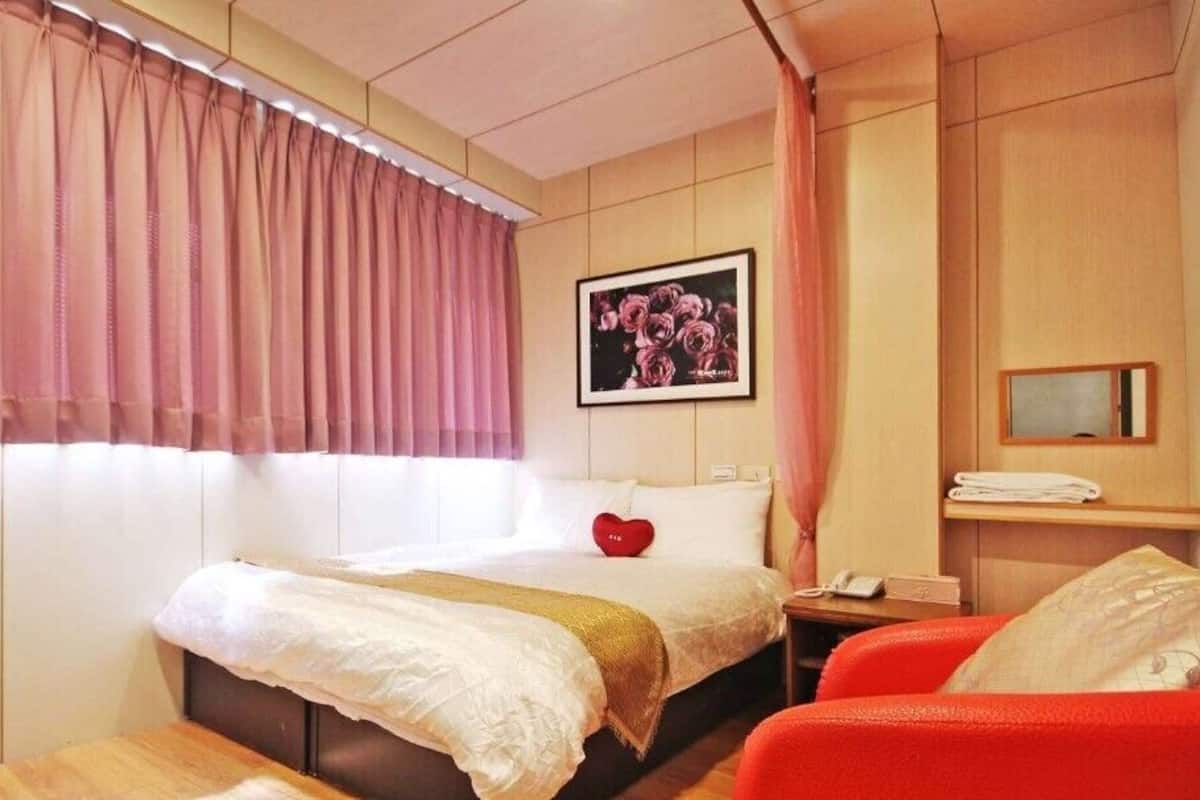 Een hotelkamer met een bed, een nachtkastje met een telefoon, een rood hartvormig kussen en een ingelijst schilderij aan de muur.