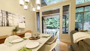 A dining area with a table set for four, a chandelier, and a view of the outdoors through large windows. - Island Beach House at Rathtrevor, Tucked in the forest and steps to the beach (Parksville)