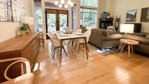 A spacious living area with a dining table, sofa, and a TV. Large windows provide natural light and a view of the outdoors. - Island Beach House at Rathtrevor, Tucked in the forest and steps to the beach (Parksville)