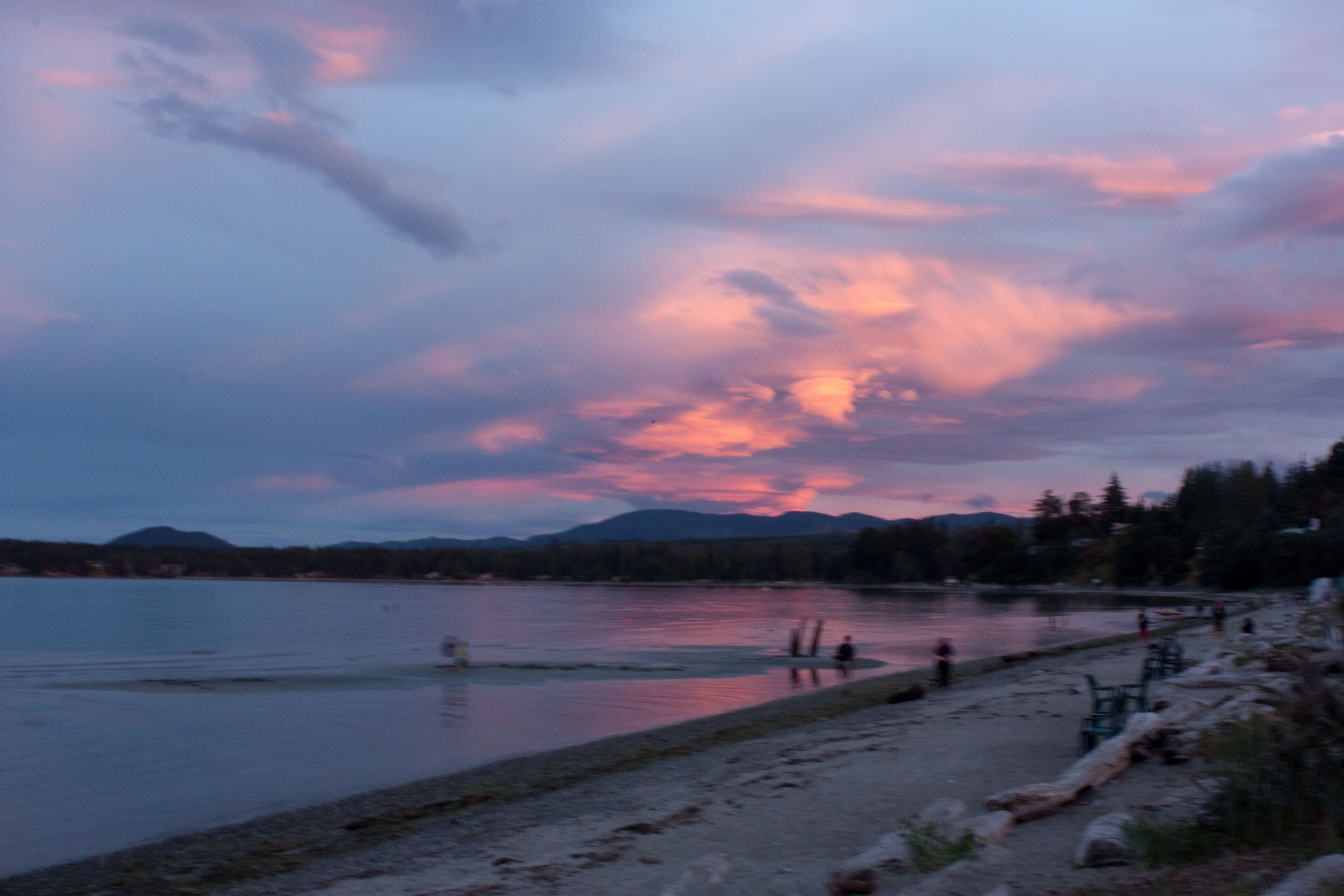 A lakeside view with a sunset, calm water, and a sandy beach with people.