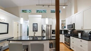 A modern kitchen with white cabinets, stainless steel appliances, a black countertop, and two bar stools. - Island Beach House at Rathtrevor, Tucked in the forest and steps to the beach (Parksville)