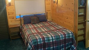 A cozy bedroom with a plaid bedspread, a window with blinds, and a wooden bookshelf. - Kenai riverfront log cabin, perfect for fishing, close to town (Sterling)