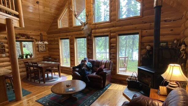 A log cabin interior with a dining area, a fireplace, and a large window overlooking a forest. - Kenai riverfront log cabin, perfect for fishing, close to town (Sterling)