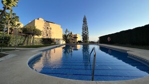 A swimming pool with clear blue water, surrounded by a paved deck. There are palm trees and a tall evergreen tree in the background. - 2-Bed Apt with Sea and views near Marbella, Spain (Calahonda)