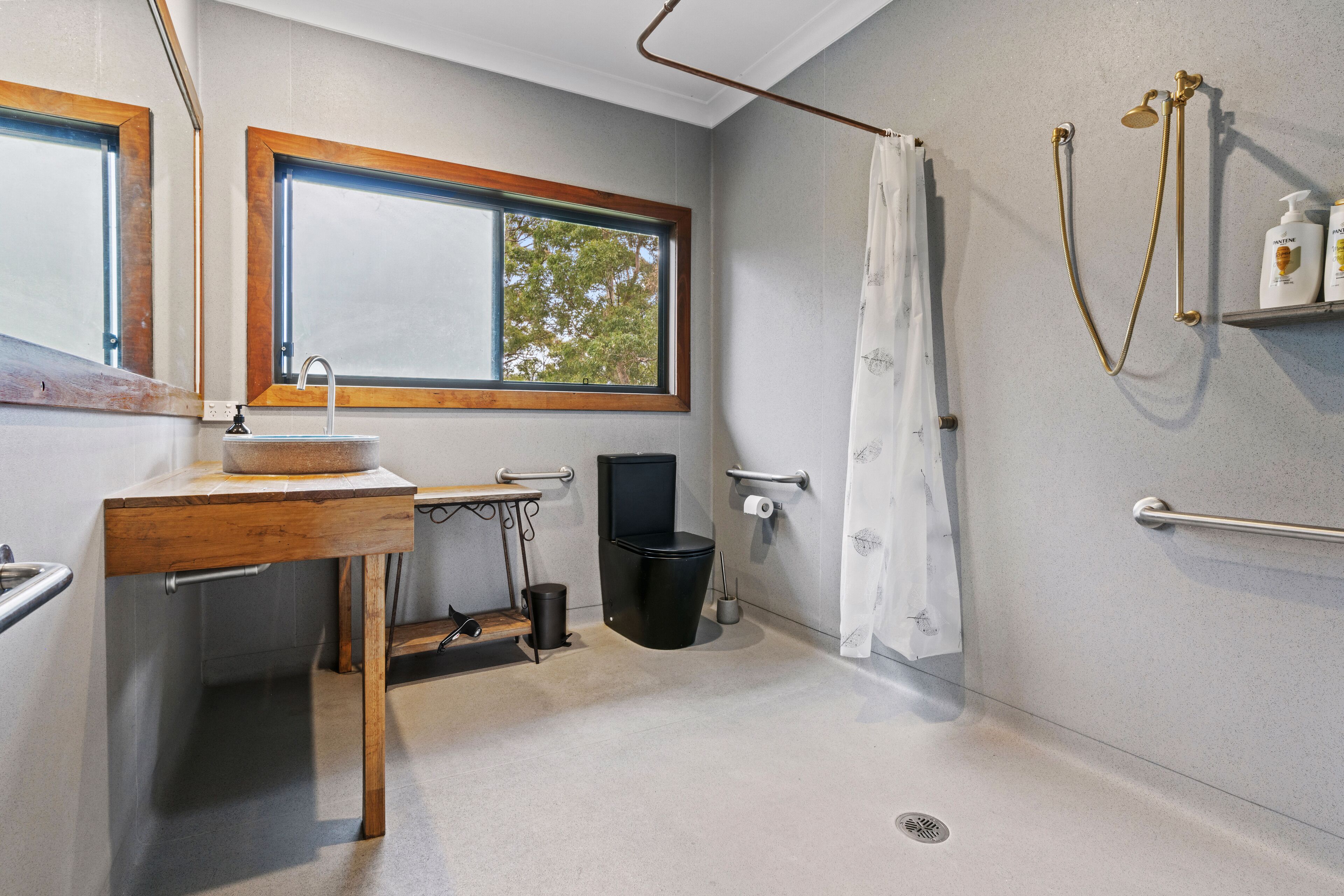 A bathroom with a wooden vanity, a window, a shower with a curtain, and a shelf with toiletries.