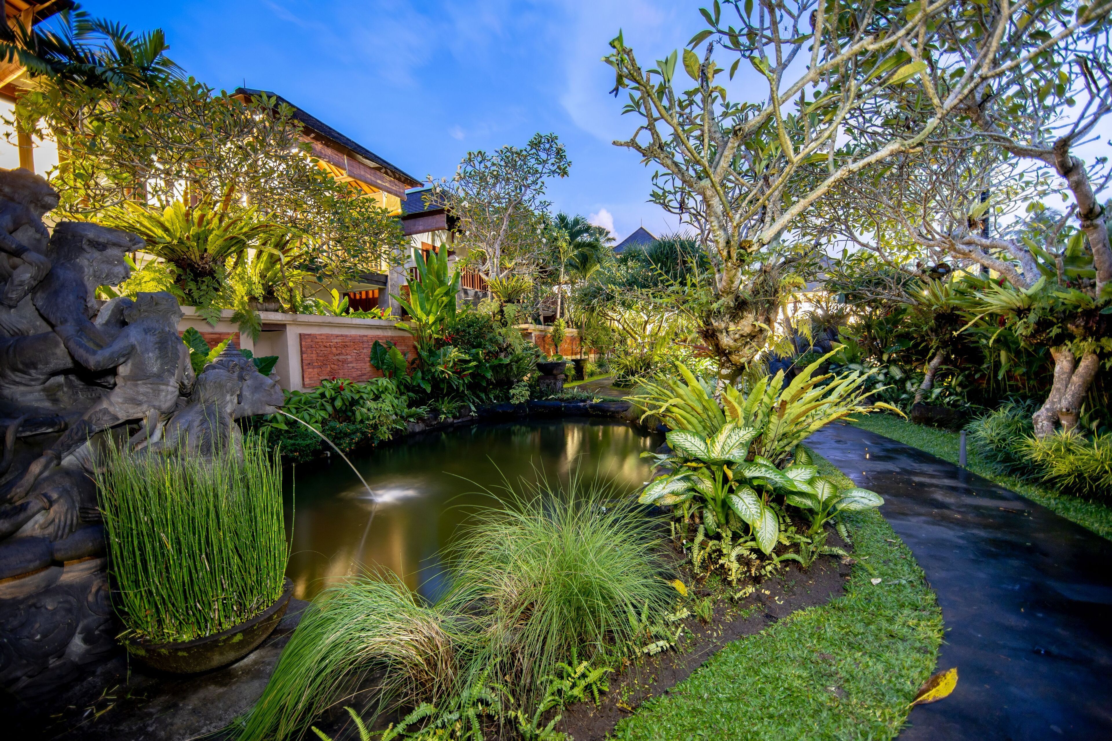 Um jardim com lago, esculturas de pedra e plantas tropicais.