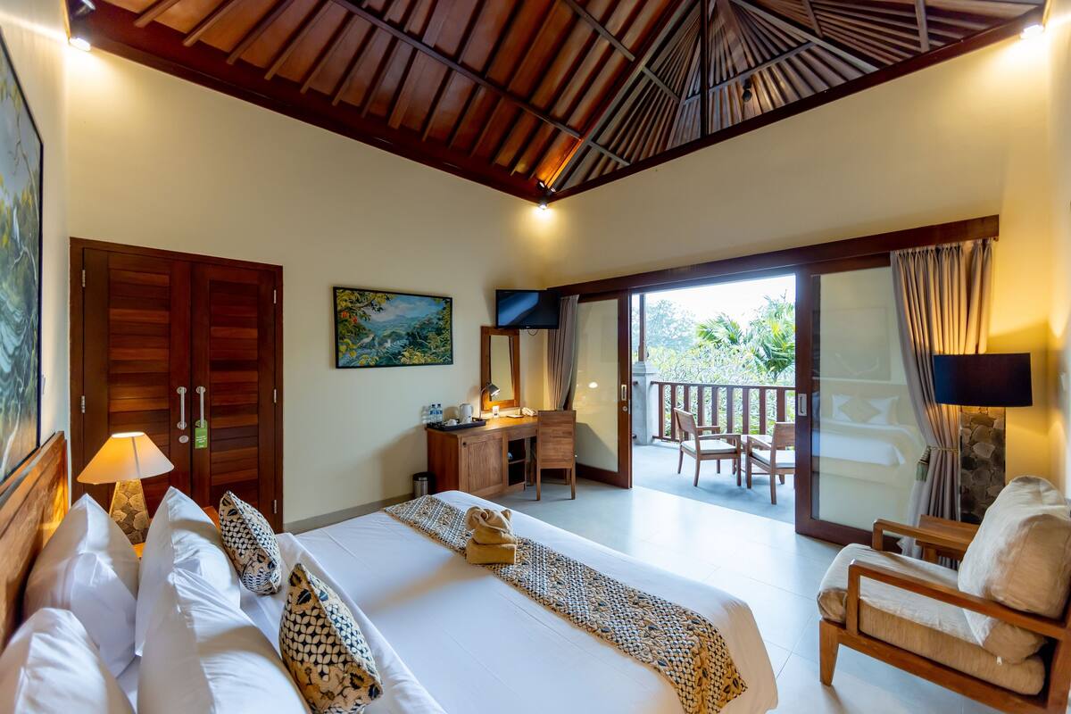 A hotel room with a bed, a desk, a chair, and a balcony with a view of greenery.