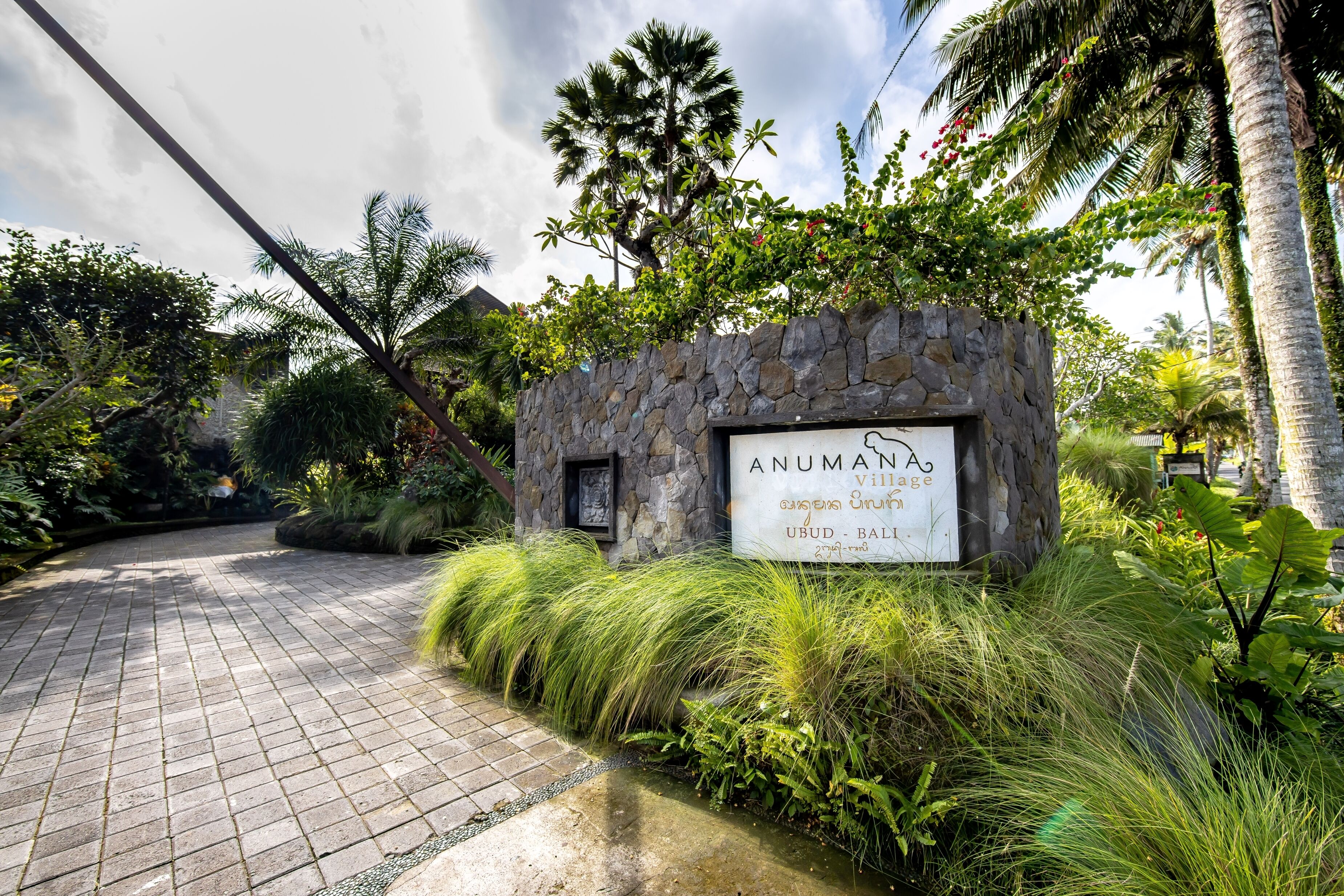 A stone sign with "Anumanah Village" and "Gunasri Creek" written on it, surrounded by lush greenery and a paved path.